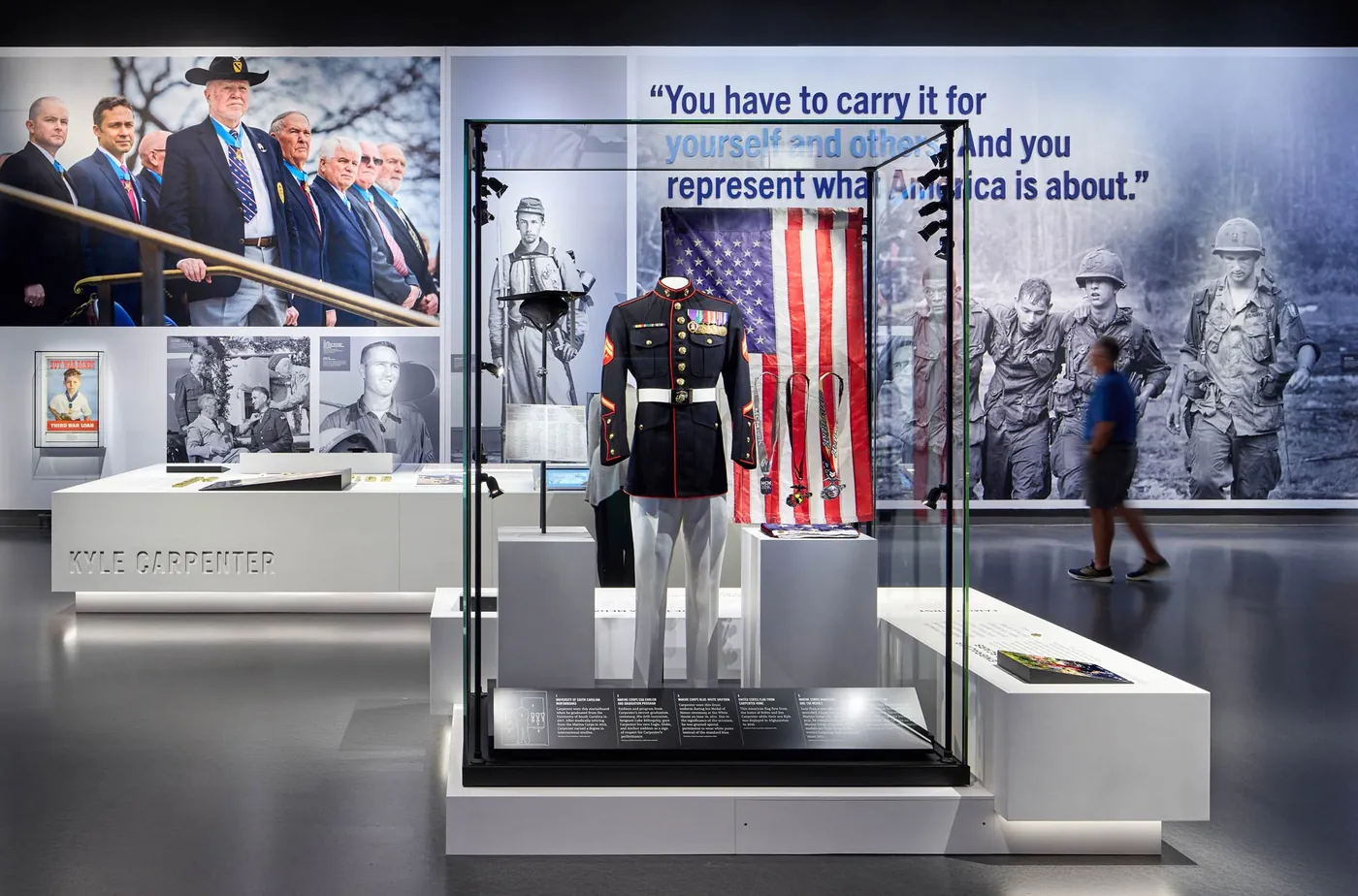 A dress uniform and flag displayed in a glass case, with a recipient's quote on the wall behind