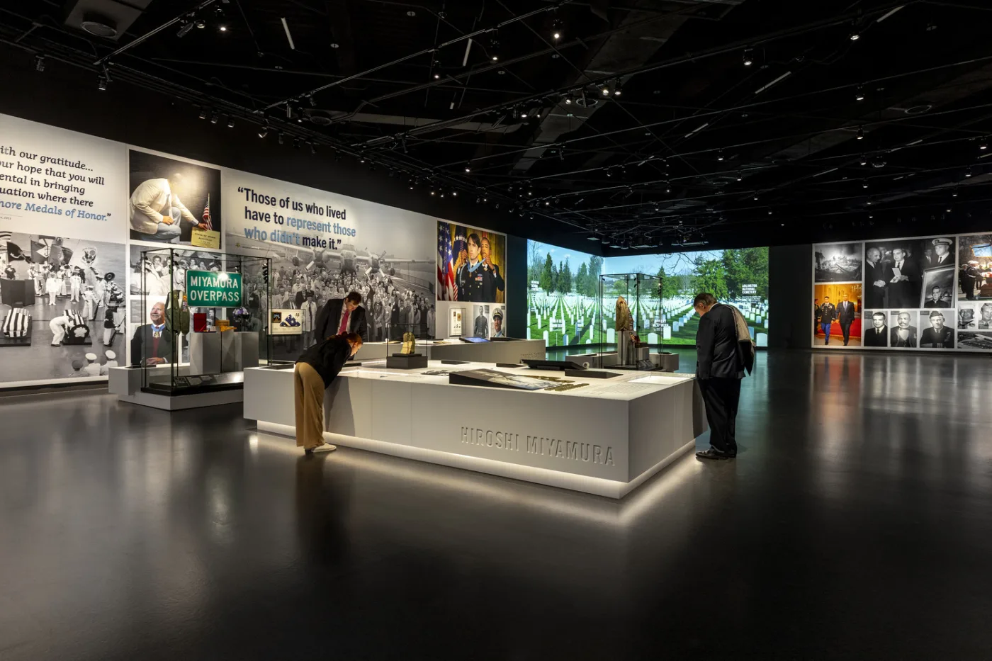 A long display table of artifacts and photographs in a dark gallery, with a large photo wall beyond