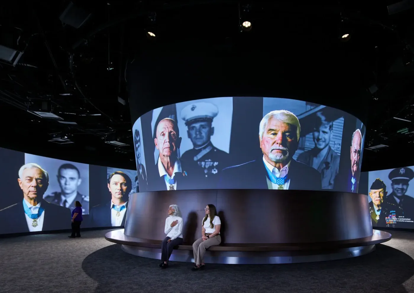 Two visitors sit beneath a circular screen of large portraits of Medal of Honor recipients