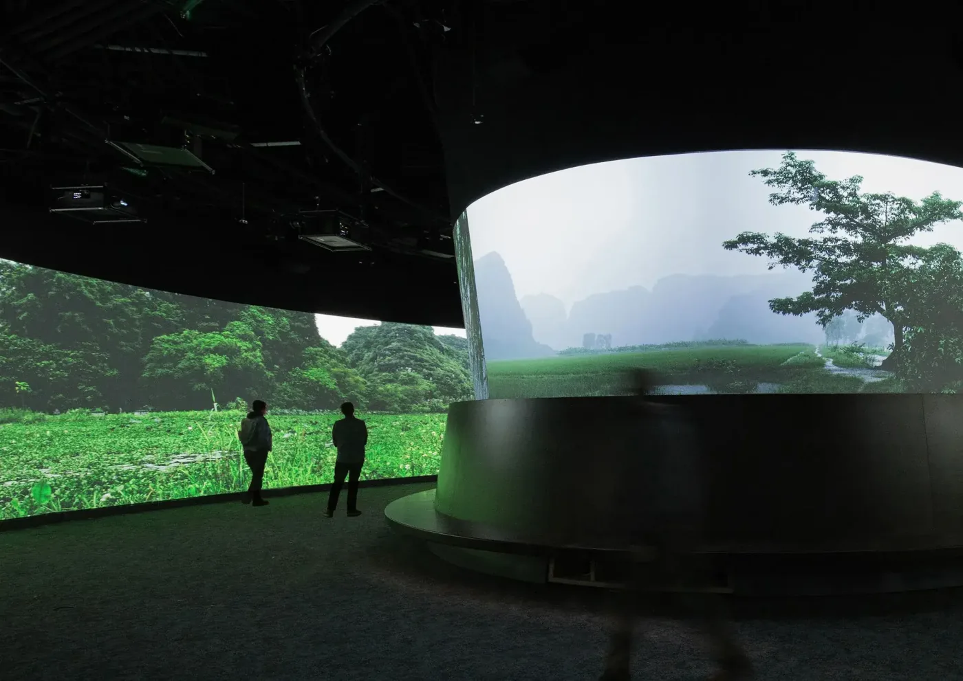 Visitors walk past a curved projection screen showing a green, misty landscape